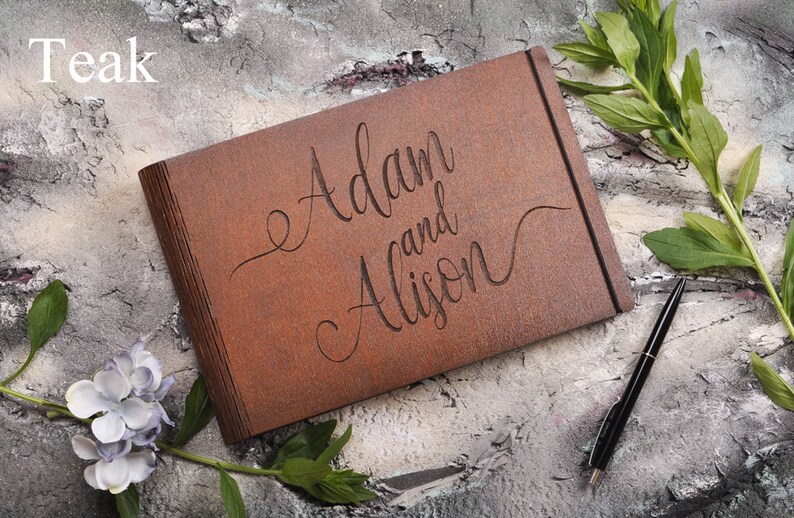 以下が含まれることがあります： 茶色の革張りゲストブックに「Adam and Alison」という名前が刻まれています。本は灰色のざらざらした表面に置かれ、ペンと緑の葉が置かれています。