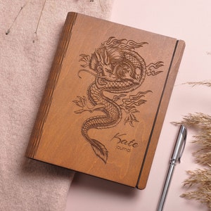 May include: A wooden journal with a dragon design engraved on the front. The journal is brown and has a black spine. The name "Kate Journal" is engraved below the dragon.