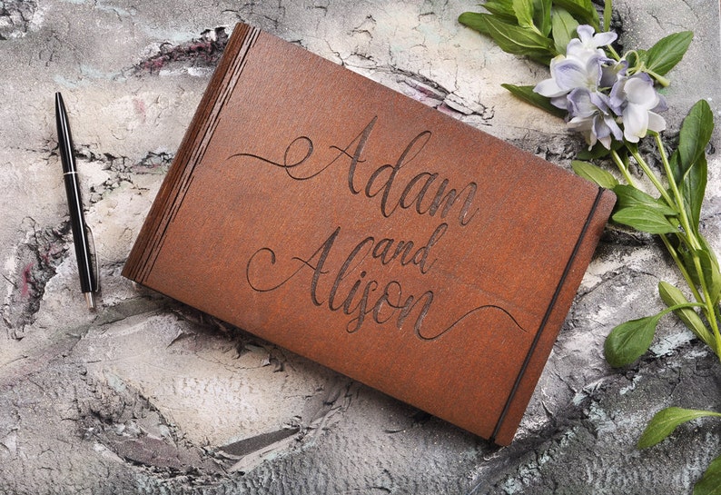 以下が含まれることがあります： 「Adam と Alison」という名前が筆記体で刻まれた茶色の木製の箱。箱は灰色のざらざらした表面に置かれ、その横に黒いペンがある。