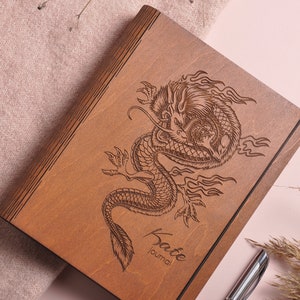 May include: A wooden journal with a dragon design engraved on the cover. The journal is brown and has a black elastic band. The text "Kate Journal" is engraved on the bottom of the cover.