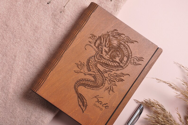 May include: A wooden journal with a dragon design engraved on the cover. The journal is brown and has a black elastic band. The text "Kate Journal" is engraved on the bottom of the cover.