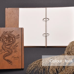 May include: A wooden journal with a dragon design engraved on the front cover. The journal is closed and has a ring binder. The journal is on a dark gray surface. The text "Kate" and "journal" is engraved on the front cover. The color of the journal is teak tree.