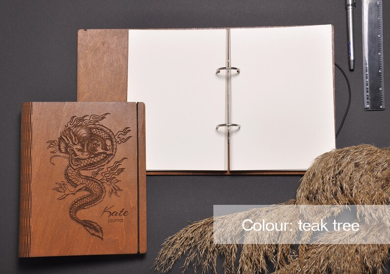 May include: A wooden journal with a dragon design engraved on the front cover. The journal is closed and has a ring binder. The journal is on a dark gray surface. The text "Kate" and "journal" is engraved on the front cover. The color of the journal is teak tree.