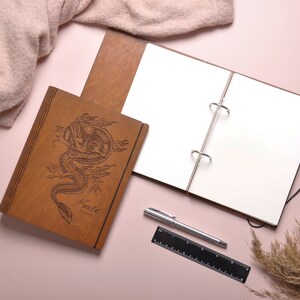 May include: A brown leather-bound journal with a dragon design and the words "Kato Journal" engraved on the cover. The journal is open to a blank page and is lying on a pink surface with a pen and a ruler.
