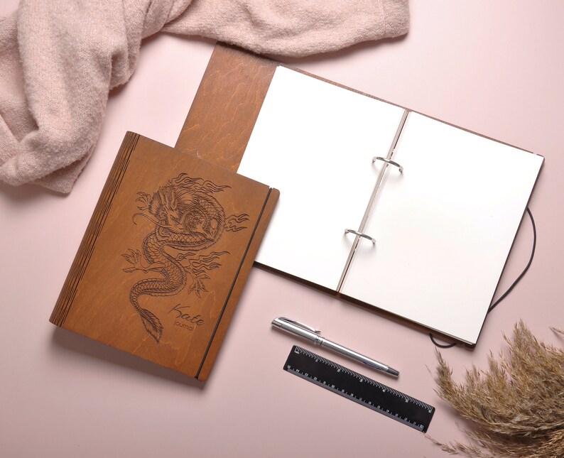 May include: A brown leather-bound journal with a dragon design and the words "Kato Journal" engraved on the cover. The journal is open to a blank page and is lying on a pink surface with a pen and a ruler.