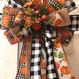 May include: A fall-themed door bow with black and white buffalo check ribbon, burlap ribbon, and orange ribbon with a pumpkin print. The bow is decorated with a cluster of faux berries and greenery.