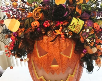 Puede incluir: Un gran recipiente con forma de calabaza naranja con una cara tallada, lleno de decoraciones de Halloween. El arreglo incluye rosas negras, caramelos de maíz, adornos brillantes y cintas. La combinación de colores es naranja, negro, morado y verde.