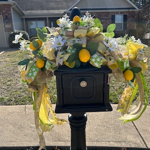 May include: A black mailbox decorated with a lemon and daisy themed wreath. The wreath features yellow and green ribbon, burlap, and artificial lemons and daisies.