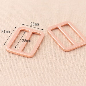 Op de afbeelding: Twee schuifgespen van roosgoudkleurig metaal, elk 25 mm breed en 31 mm lang.