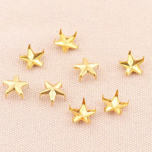 May include: Eight gold star-shaped metal studs with pointed tips. The studs are arranged in a scattered pattern on a light pink fabric background.