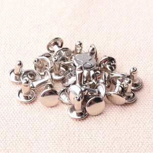 May include: A pile of silver metal rivets, also known as studs, for leather craft projects. The rivets have a round head and a pointed post.