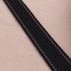 May include: Close-up of a black strap with reflective stitching. The strap is on a light brown fabric background.