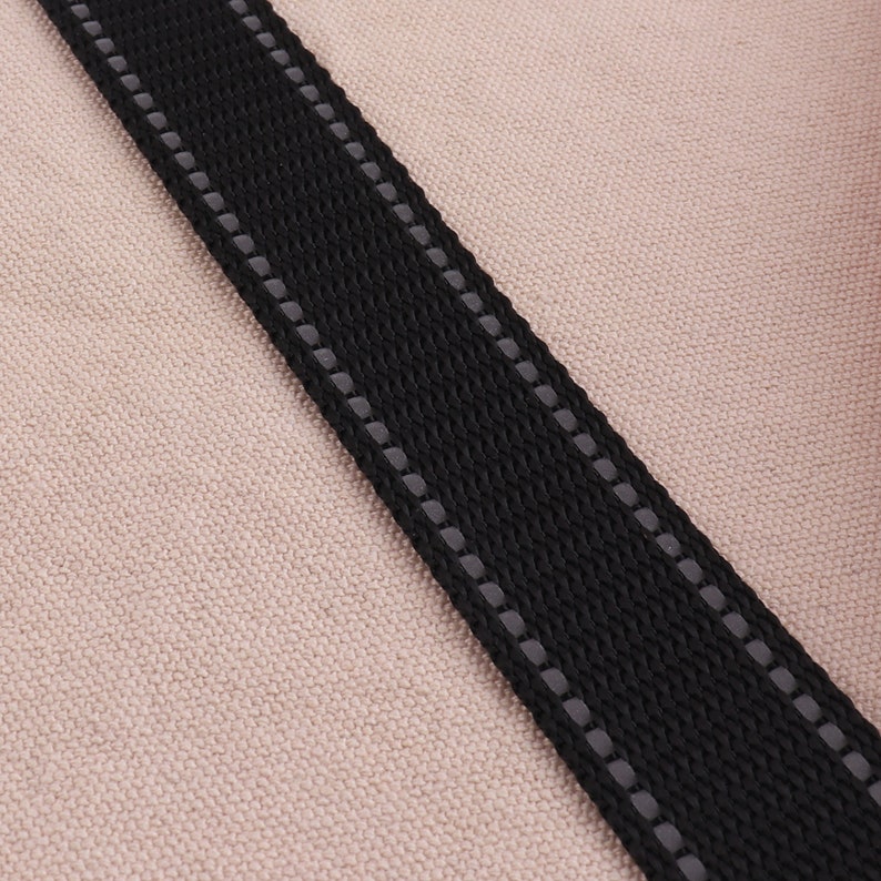 May include: Close-up of a black strap with reflective stitching. The strap is on a light brown fabric background.