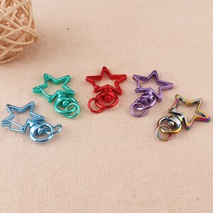 Puede incluir: Cinco llaveros con forma de estrella en diferentes colores: verde azulado, rojo, morado, dorado e iridiscente. Cada llavero tiene un anillo giratorio.