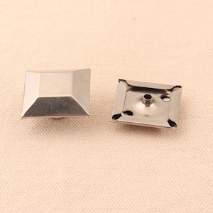 May include: Two silver metal square-shaped rivets with a raised center. One rivet has a hole in the center and the other rivet is flat.