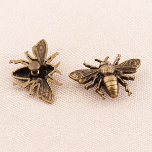 Puede incluir: Dos botones de metal en forma de abeja de color dorado antiguo. Un botón es una abeja plana con las alas extendidas. El otro botón es una abeja con las alas plegadas y un poste en la parte posterior para coser.