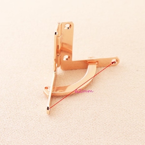 Può includere: Cerniera in metallo dorato con una misura di 62 mm segnata sull'immagine. La cerniera è progettata per piegarsi ad un angolo di 90 gradi.