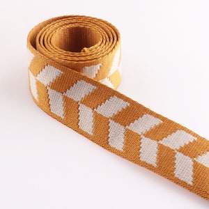 May include: A close-up of a mustard yellow woven ribbon with a white geometric pattern. The ribbon is rolled up and the pattern is visible on the front and back of the ribbon.