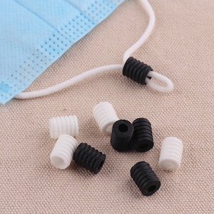 May include: A close-up of a group of black and white silicone cord stoppers. The stoppers are designed to keep cords from fraying and are a popular accessory for face masks.