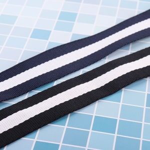 May include: Two striped fabric straps, one navy blue with a white stripe and one black with a white stripe. The straps are lying on a blue tiled surface.
