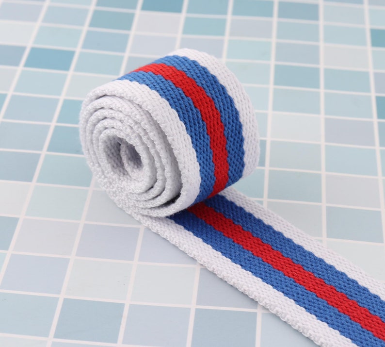 K&ouml;nnte beinhalten: Wei&szlig;es Baumwollband mit roten und blauen Streifen. Das Band ist aufgerollt und die Streifen sind sichtbar.
