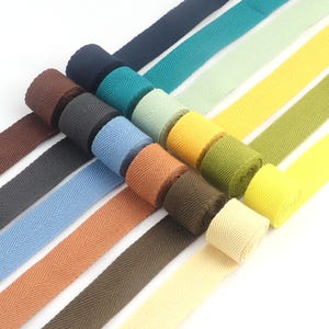 May include: A collection of colorful herringbone twill tapes in various shades, including blue, green, yellow, and brown. The tapes are rolled and unrolled, showcasing their texture and color variations. Ideal for sewing and crafting projects.