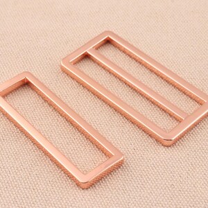 May include: Two rose gold metal belt buckle slides. The slides are rectangular with a smooth, polished finish.