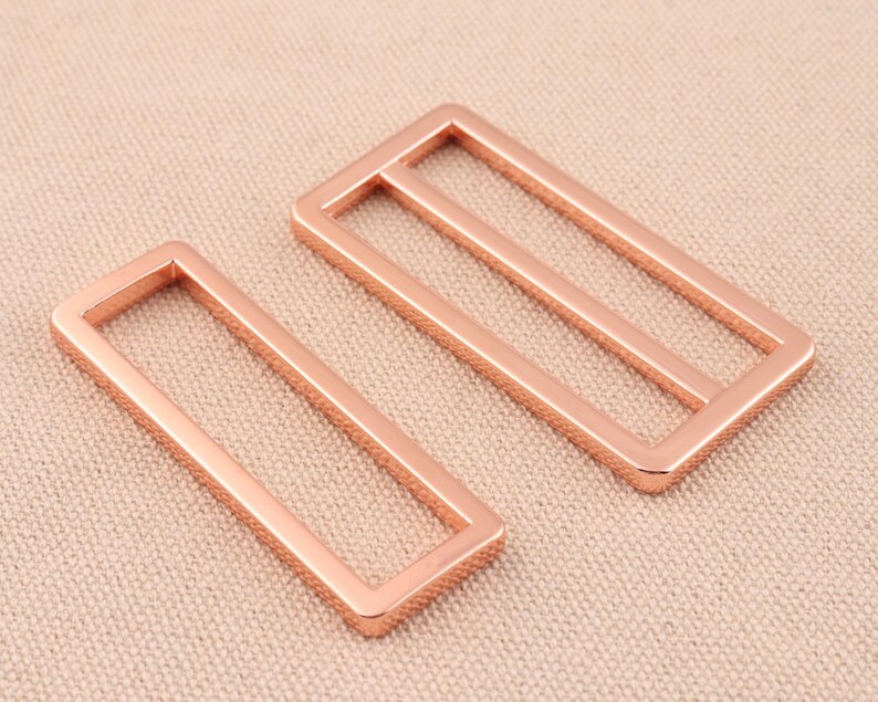 May include: Two rose gold metal belt buckle slides. The slides are rectangular with a smooth, polished finish.
