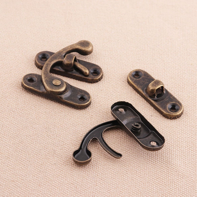 May include: Four antique bronze-colored metal latches. Two are assembled, and two are disassembled. Each latch has a curved hook and a rectangular base with screw holes. The metal has a distressed finish.