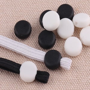 Pode incluir: Close-up de tampas redondas de silicone preto e branco para máscaras faciais. As tampas são projetadas para ajudar a manter as correias da máscara no lugar.