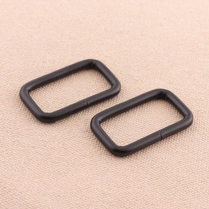 May include: Two black metal rectangular rings, also known as D-rings, are shown on a light brown fabric background.