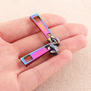 May include: Two iridescent zipper pulls with a rectangular shape and a silver metal slider. The zipper pulls have a rainbow color effect.