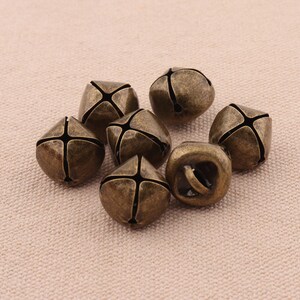 May include: A collection of seven antique bronze-colored jingle bells. Each bell has a cross-shaped design on its sides and a loop for hanging. The bells are clustered together on a neutral-toned fabric surface.