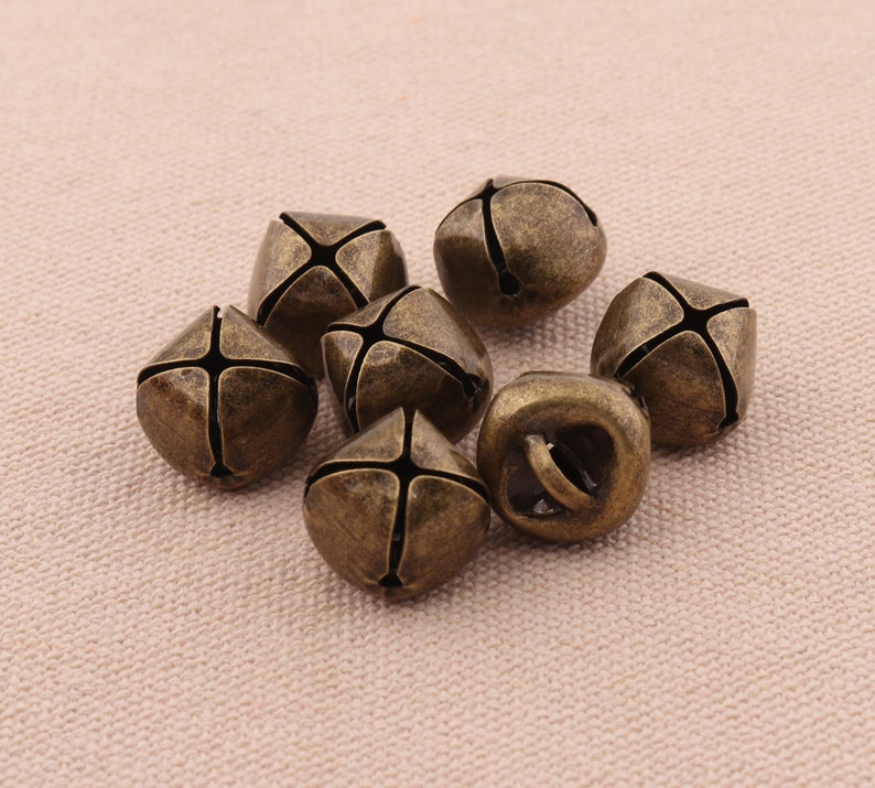May include: A collection of seven antique bronze-colored jingle bells. Each bell has a cross-shaped design on its sides and a loop for hanging. The bells are clustered together on a neutral-toned fabric surface.