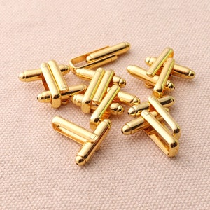 May include: A group of gold-toned cufflinks with a geometric design. The cufflinks are scattered on a light beige fabric surface.