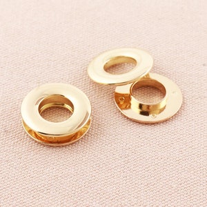 May include: Three gold metal eyelets, each with a circular hole in the center. The eyelets are arranged in a staggered pattern on a light pink fabric surface.