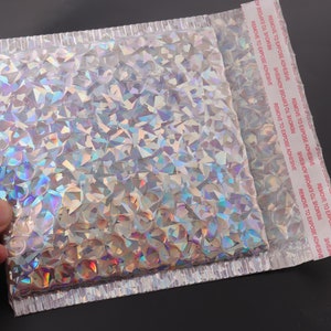 Op de afbeelding: Een zilveren, iriserende, gevoerde envelop met een rood "REMOVE TO EXPOSE ADHESIVE"-label aan de zijkant.