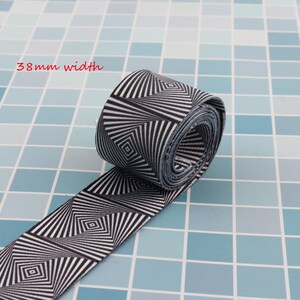 May include: Black and white elastic band with a geometric pattern. The band is 38 mm wide and is rolled up on a blue tiled surface.