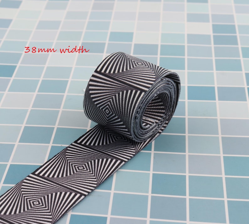 May include: Black and white elastic band with a geometric pattern. The band is 38 mm wide and is rolled up on a blue tiled surface.