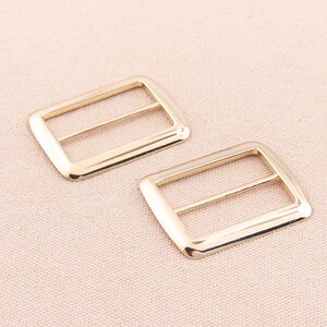 May include: Two gold-toned metal slider buckles. The buckles are rectangular with rounded corners and have two parallel slots for a strap.