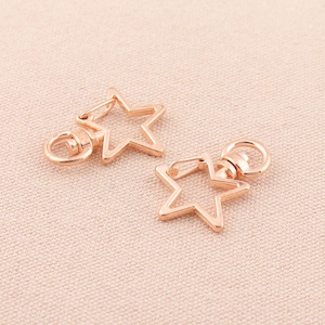 May include: Two gold-toned metal star-shaped keyrings with a swivel clasp. The keyrings are on a light brown fabric background.