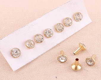 May include: A set of gold-toned metal studs with clear rhinestone accents. The studs are arranged on a white card and some are shown individually on a beige surface.