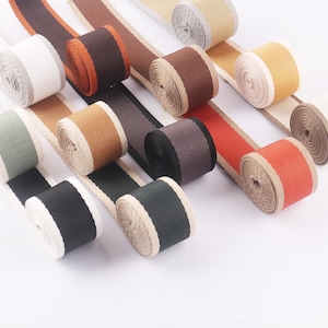 Può includere: Una collezione di rotoli di nastro colorati in varie tonalità di marrone, arancione, nero e verde. Ogni rotolo ha un bordo a contrasto e i nastri sono disposti su una superficie bianca. Ideale per progetti di artigianato e decorativi.