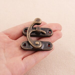 May include: An antique bronze-colored clasp, featuring a curved hook and two rectangular plates with screw holes. The clasp is designed for securing boxes or small containers, showcasing a vintage aesthetic.