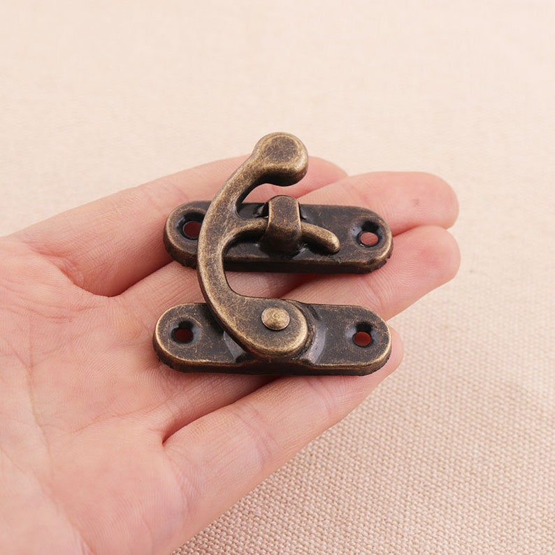 May include: An antique bronze-colored clasp, featuring a curved hook and two rectangular plates with screw holes. The clasp is designed for securing boxes or small containers, showcasing a vintage aesthetic.