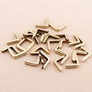 May include: A pile of gold-colored metal corner protectors. The corner protectors are shaped like an inverted V and have a smooth, shiny finish.
