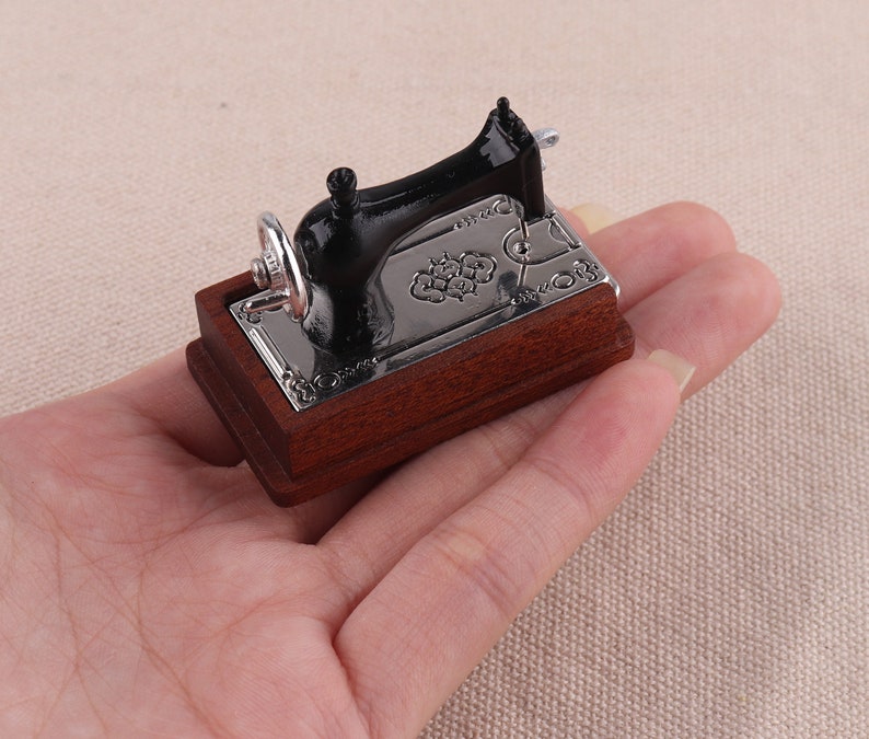 May include: A miniature black sewing machine with a wooden base. The machine has a silver metal plate with an ornate design.