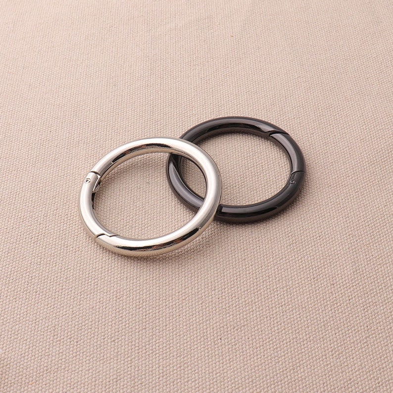 May include: Two metal rings, one silver and one black, with a snap closure. The rings are on a beige fabric background.