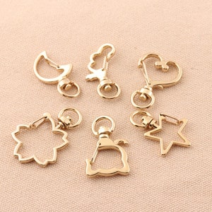 May include: A set of seven gold-toned metal keychains with different shapes, including a crescent moon, a star, a heart, a flower, and a cat. The keychains are all attached to a swivel clasp.