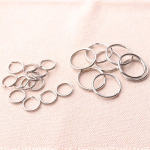May include: A close-up of a pile of silver key rings. The key rings are arranged in a random pattern on a light brown fabric surface.